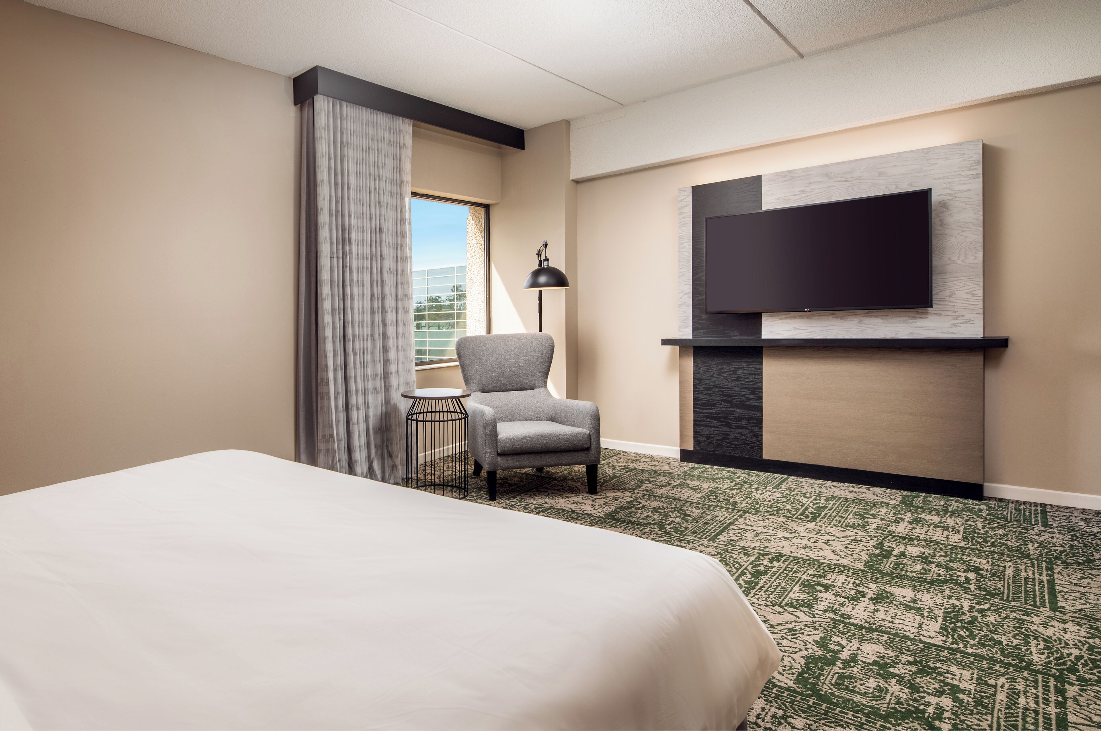 A hotel room with a large bed, a grey armchair, a small side table, a flat-screen TV mounted on the wall, and a patterned carpet.