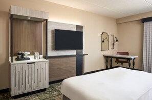 Una habitación de hotel con cama, televisor montado en la pared, escritorio con silla y un minibar.