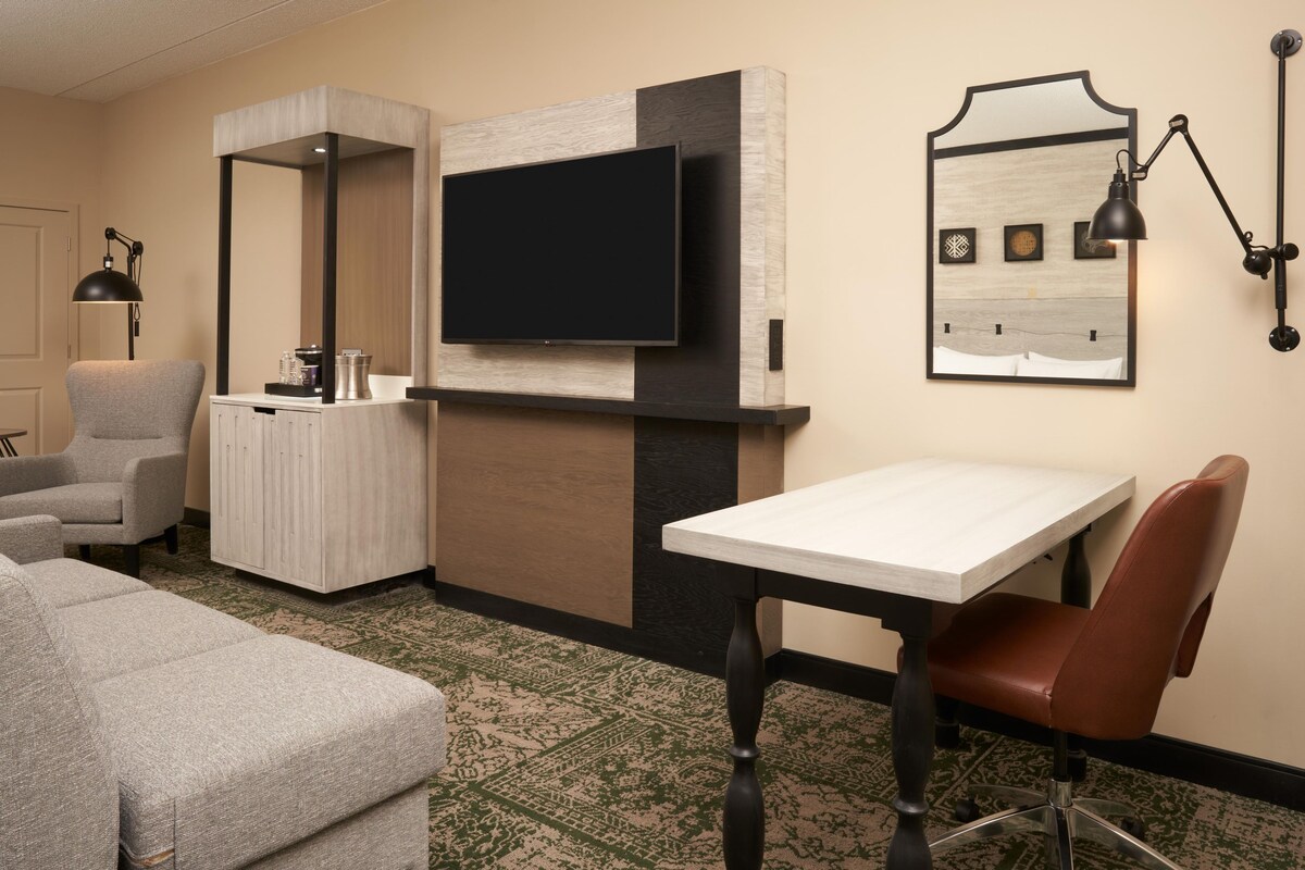 A hotel room with a flat-screen TV mounted on a wall, a small table with a chair, a sofa, and a cabinet with a mirror.