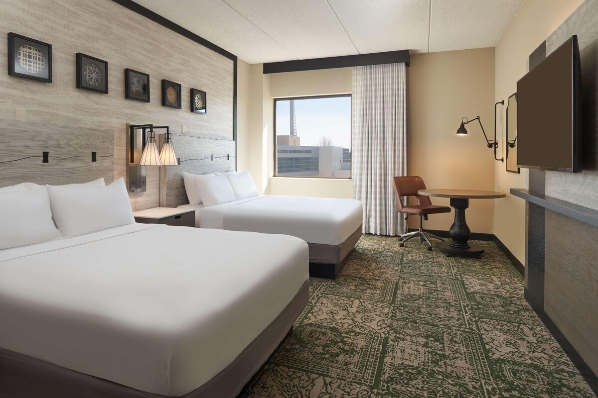 A hotel room with two beds, a desk, a chair, a TV, and a window with a city view.