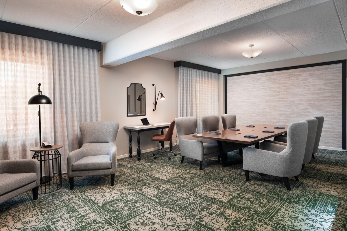 A conference room with a round table, six chairs, a laptop, a desk lamp, and a patterned carpet.