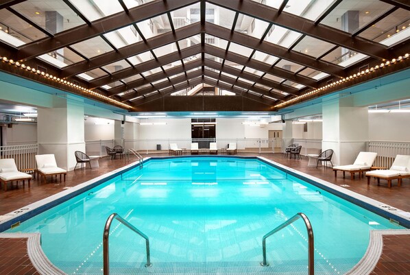 Una piscina interior con agua cristalina y azul, rodeada de sillones de descanso y un techo alto con tragaluces.