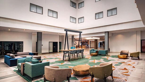 A spacious lobby with a central table and chairs, surrounded by sofas and ottomans, and large windows with curtains. - Hilton Appleton Paper Valley (Appleton)