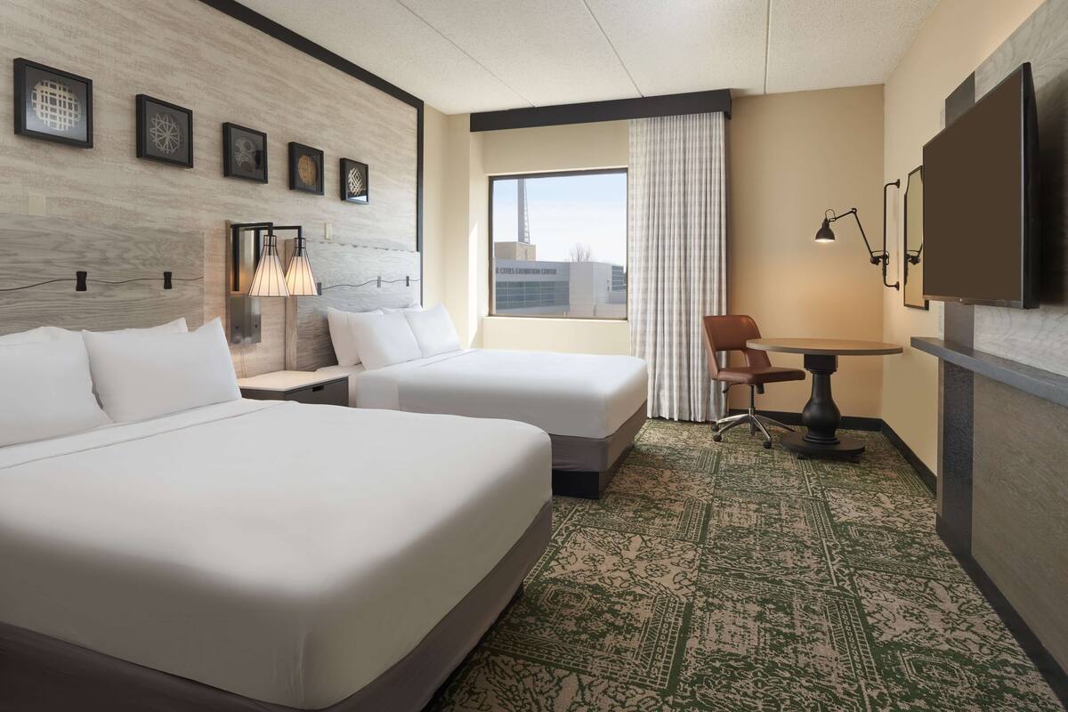 A hotel room with two beds, a desk, a chair, a TV, and a window with a city view.