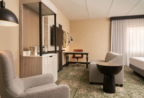A hotel room with a sofa, a chair, a small table, a lamp, a TV, and a window with curtains. - Hilton Appleton Paper Valley (Appleton)