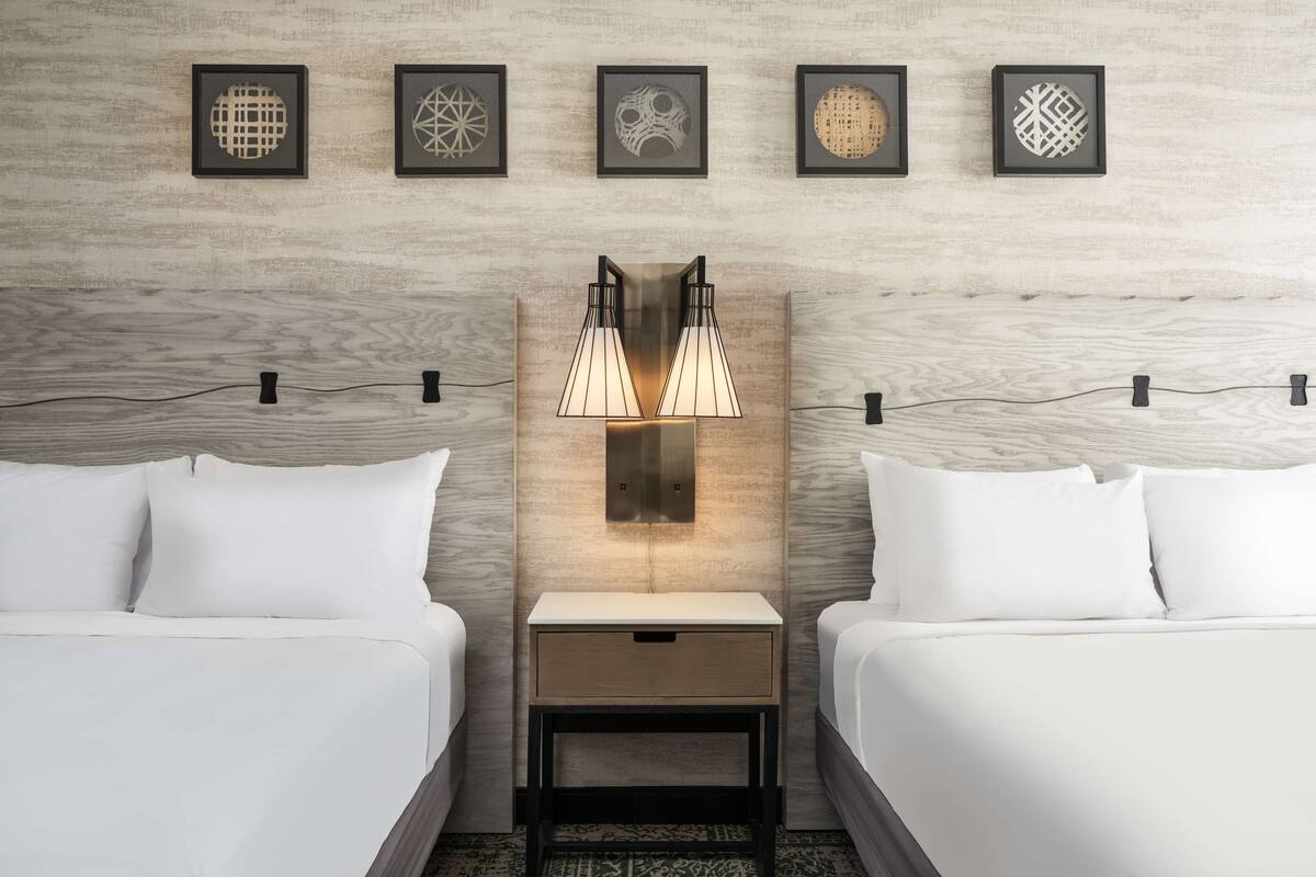 A hotel room with two beds, a nightstand with a lamp, and four framed wall art pieces.