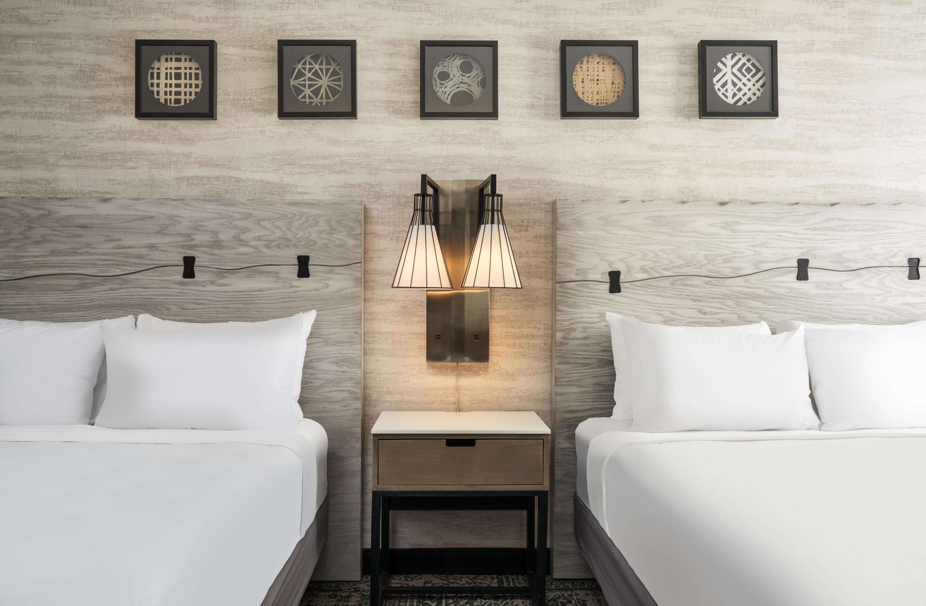 Habitación de hotel con dos camas, una mesita de noche, una lámpara de pared y cinco cuadros enmarcados.