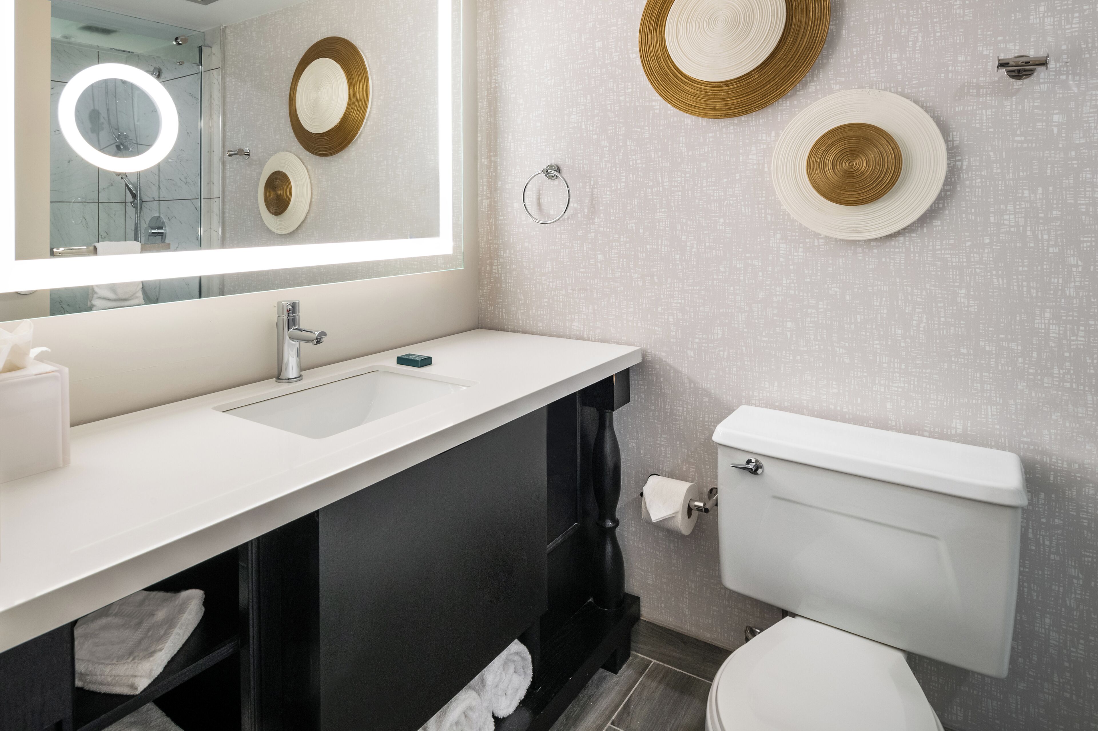 Ein modernes Badezimmer mit einem weißen Waschbecken, einer schwarzen Kommode und einer Toilette. An der Wand befinden sich dekorative Teller und ein runder Spiegel über dem Waschbecken.