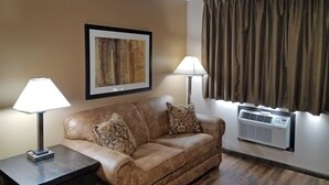 A room with a brown leather sofa, two patterned pillows, a framed abstract painting, two floor lamps, and an air conditioning unit. - Americas Best Value Inn & Suites Bismarck (Bismarck)