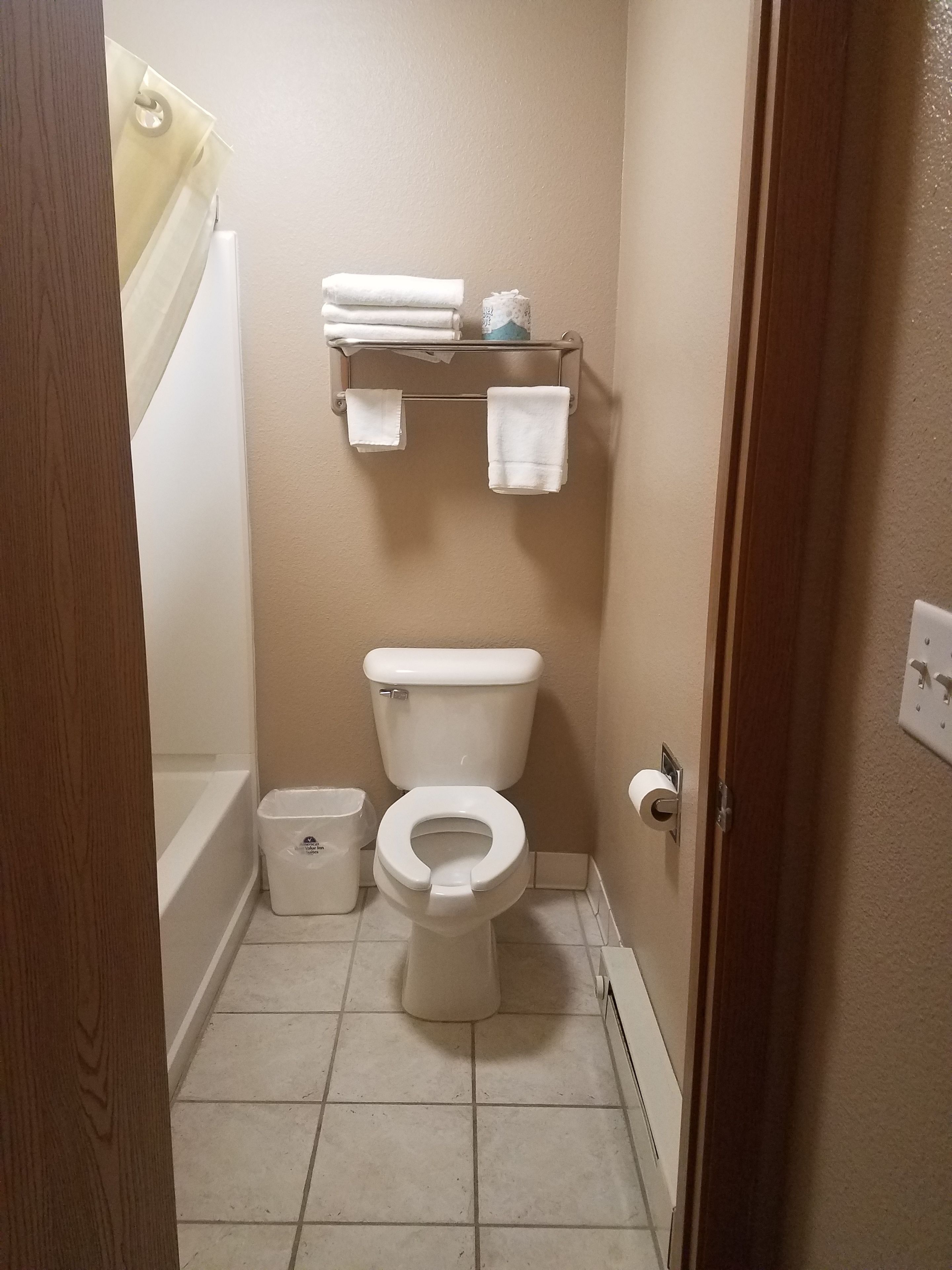 A small bathroom with a toilet, a toilet paper holder, a towel rack with folded towels, a trash bin, and a shower curtain.