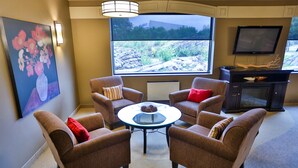 A room with four brown armchairs, a small round table, a flat-screen TV, and a large window with a view of rocks and greenery. - Travelway Inn Sudbury (Sudbury)