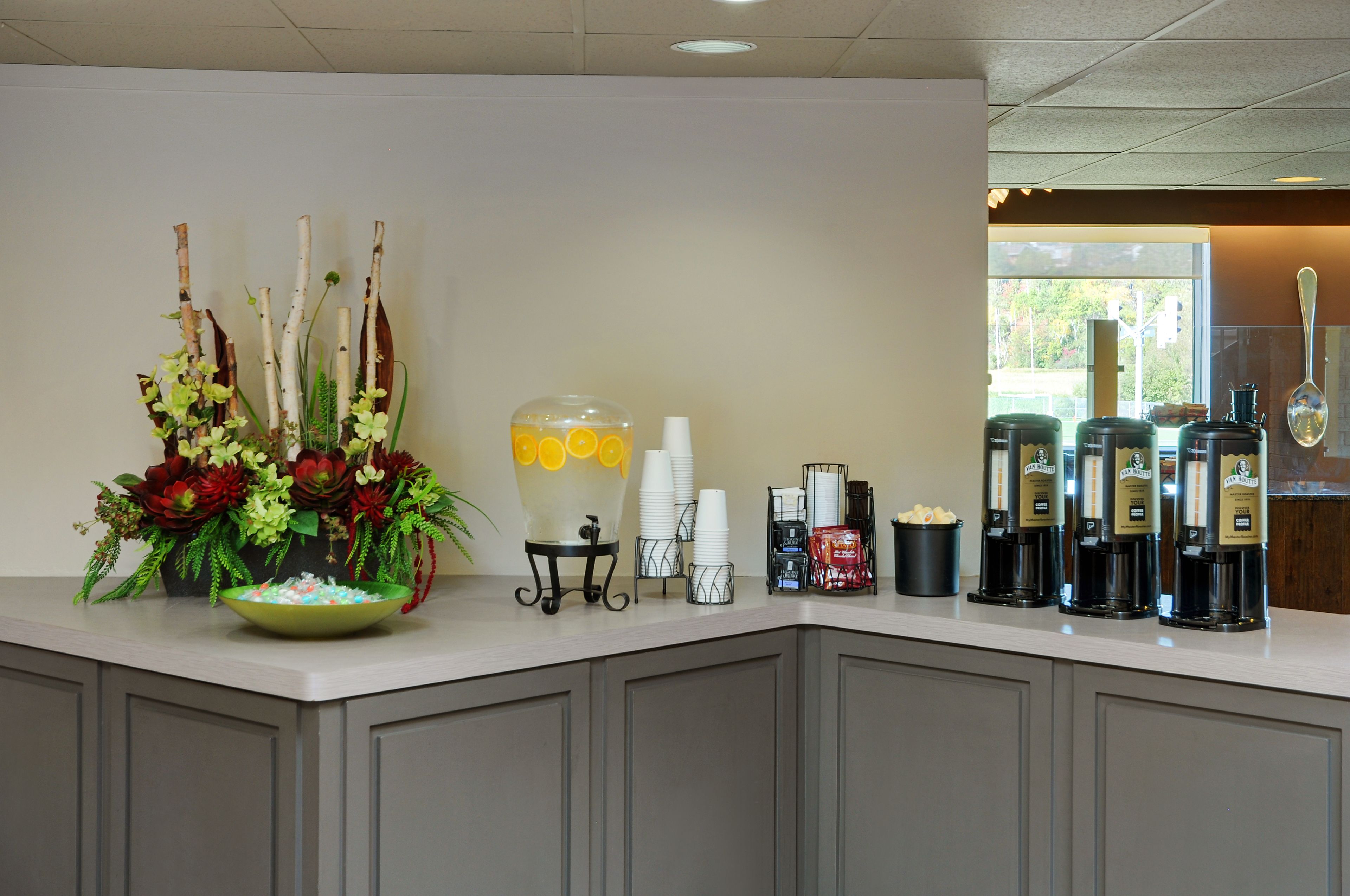 Een ontbijtbuffet met een bloemstuk, een fruitschaal, gestapelde witte bekers en koffieautomaten.