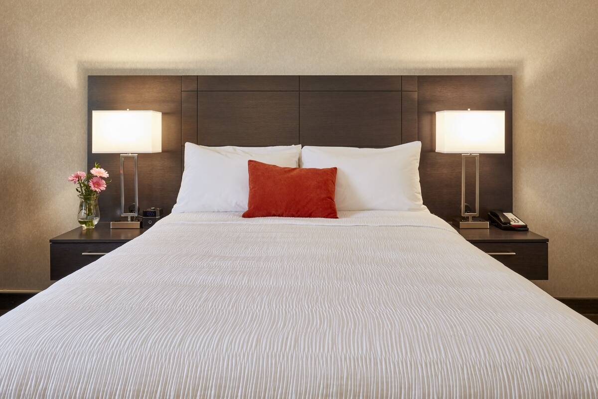 A neatly made bed with white linens and a single red accent pillow, flanked by two bedside tables with lamps and a vase of flowers.