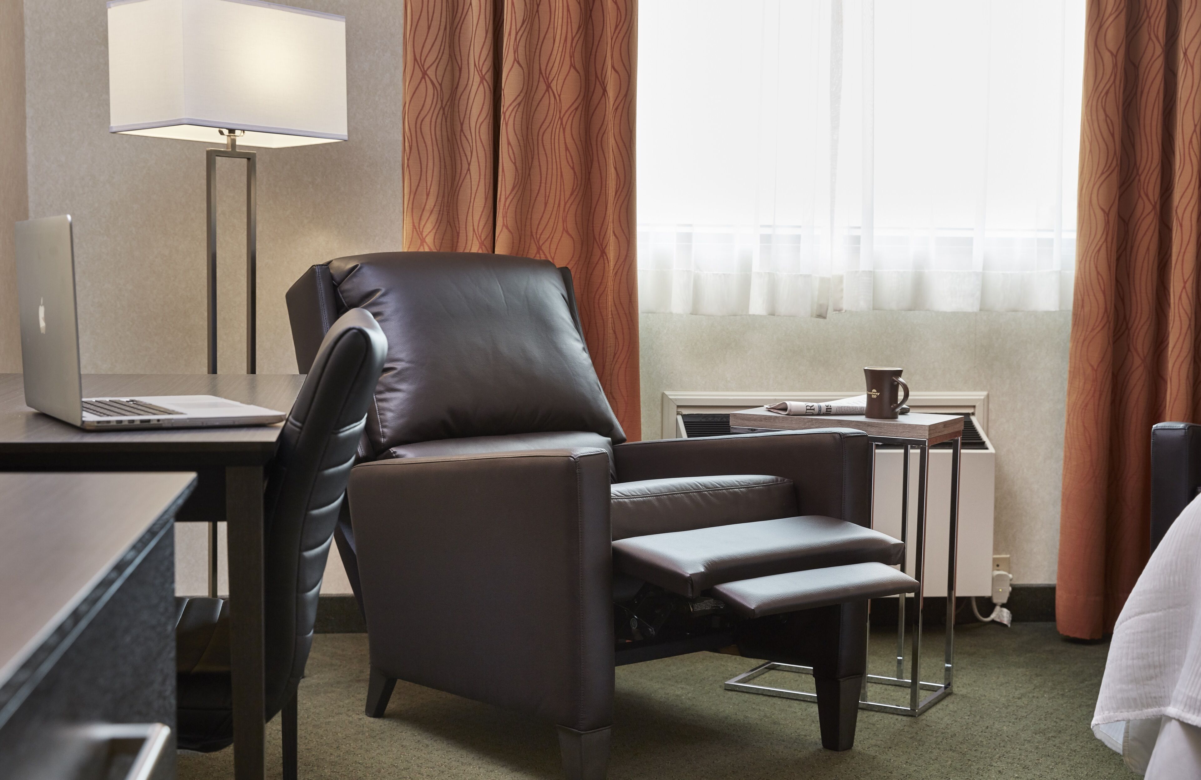 A hotel room with a leather chair, a desk with a laptop, a small table with a coffee cup, and a window with curtains.