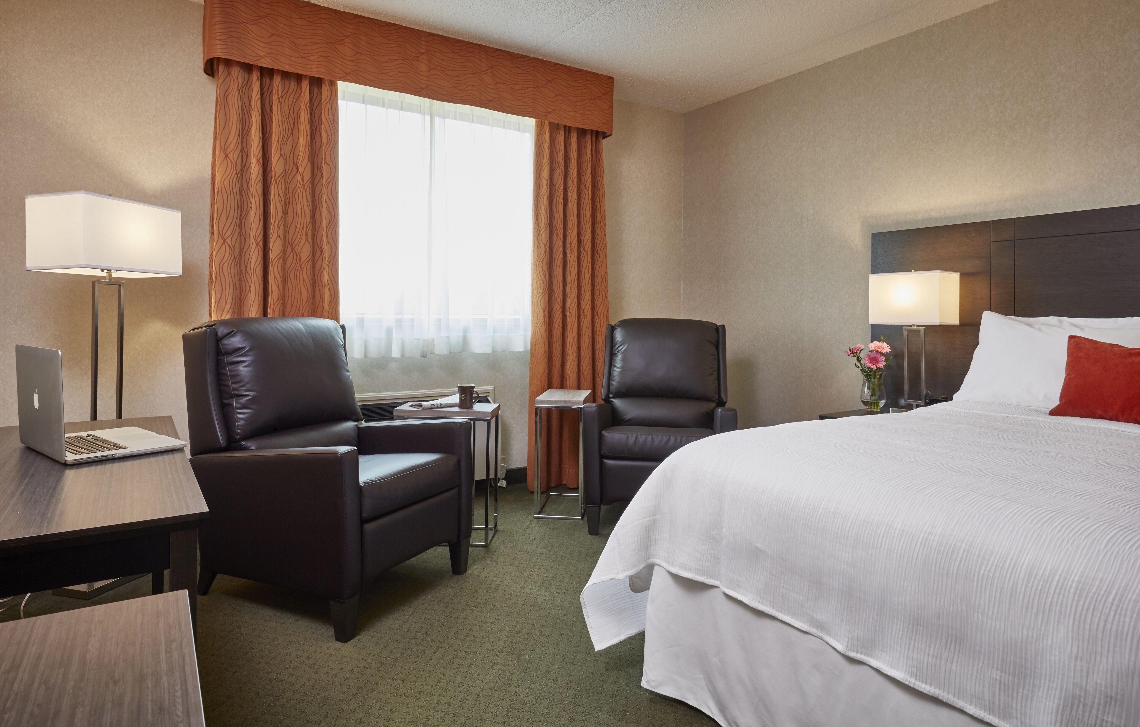 Una habitación de hotel con una cama grande, dos sillas, un escritorio con una computadora portátil, una lámpara y una ventana con cortinas.