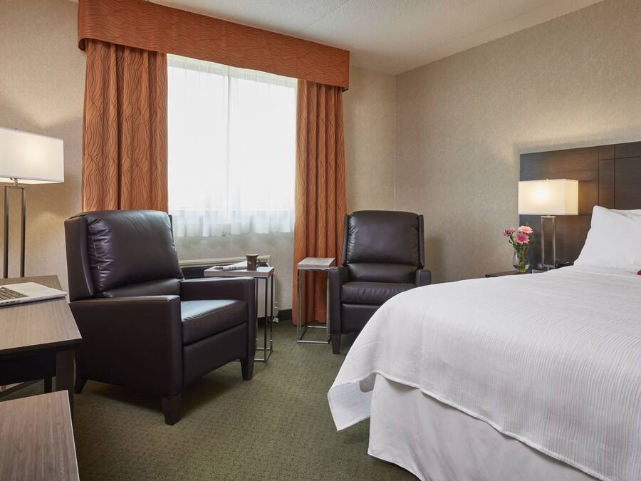 Una habitación de hotel con una cama grande, dos sillas, un escritorio con una computadora portátil, una lámpara y una ventana con cortinas.