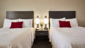A hotel room with two beds, each with white linens and red pillows, a nightstand with a lamp, and a small table with a vase of flowers. - Travelway Inn Sudbury (Sudbury)