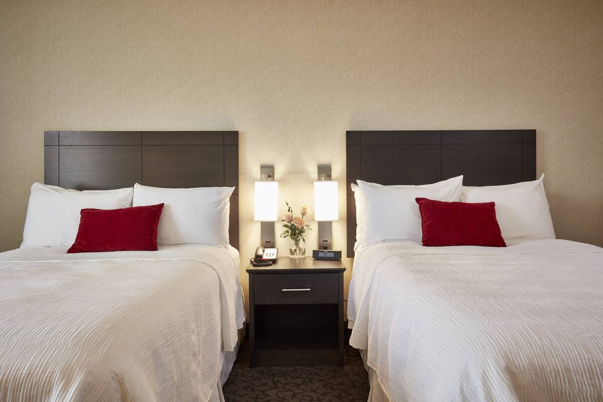 A hotel room with two beds, each with white linens and red pillows, a nightstand with a lamp, and a small table with a vase of flowers.