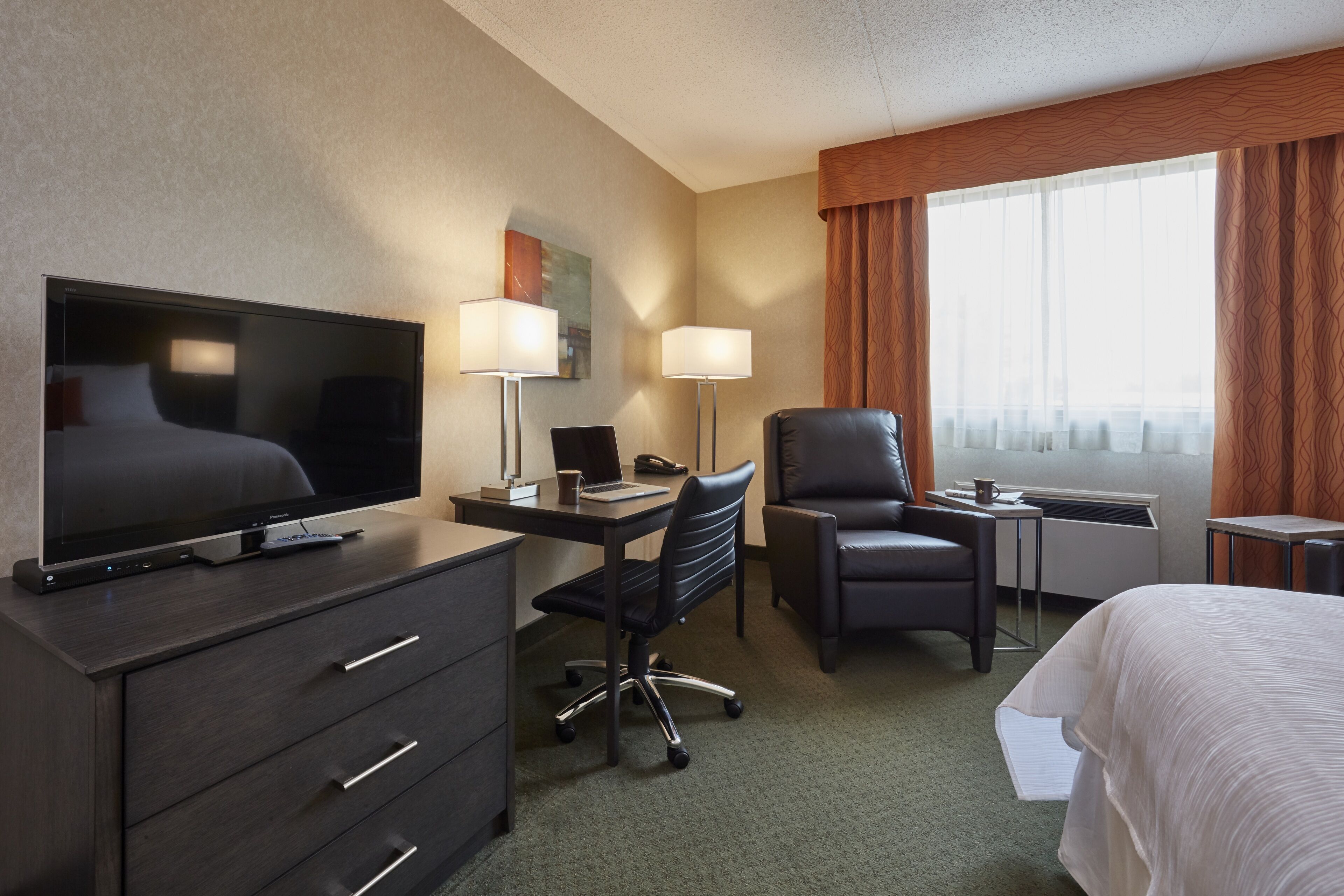 Habitación de hotel con una cama, un escritorio con computadora, un televisor, una silla y una ventana con cortinas.