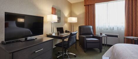 Habitación de hotel con una cama, un escritorio con computadora, un televisor, una silla y una ventana con cortinas.