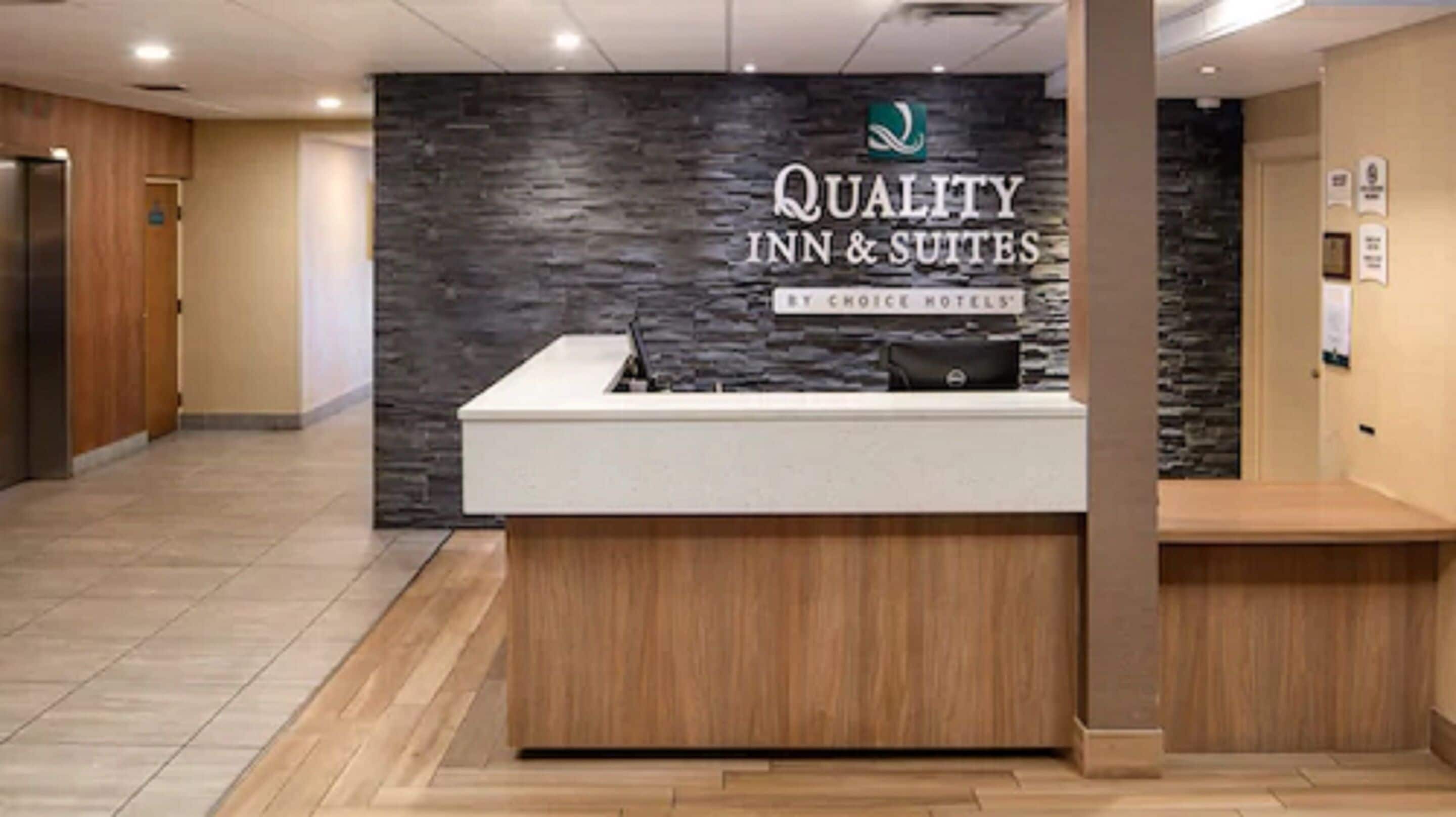 A reception desk with a stone wall backdrop, a sign reading "Quality Inn & Suites by Choice Hotels", and a black bag on the counter.