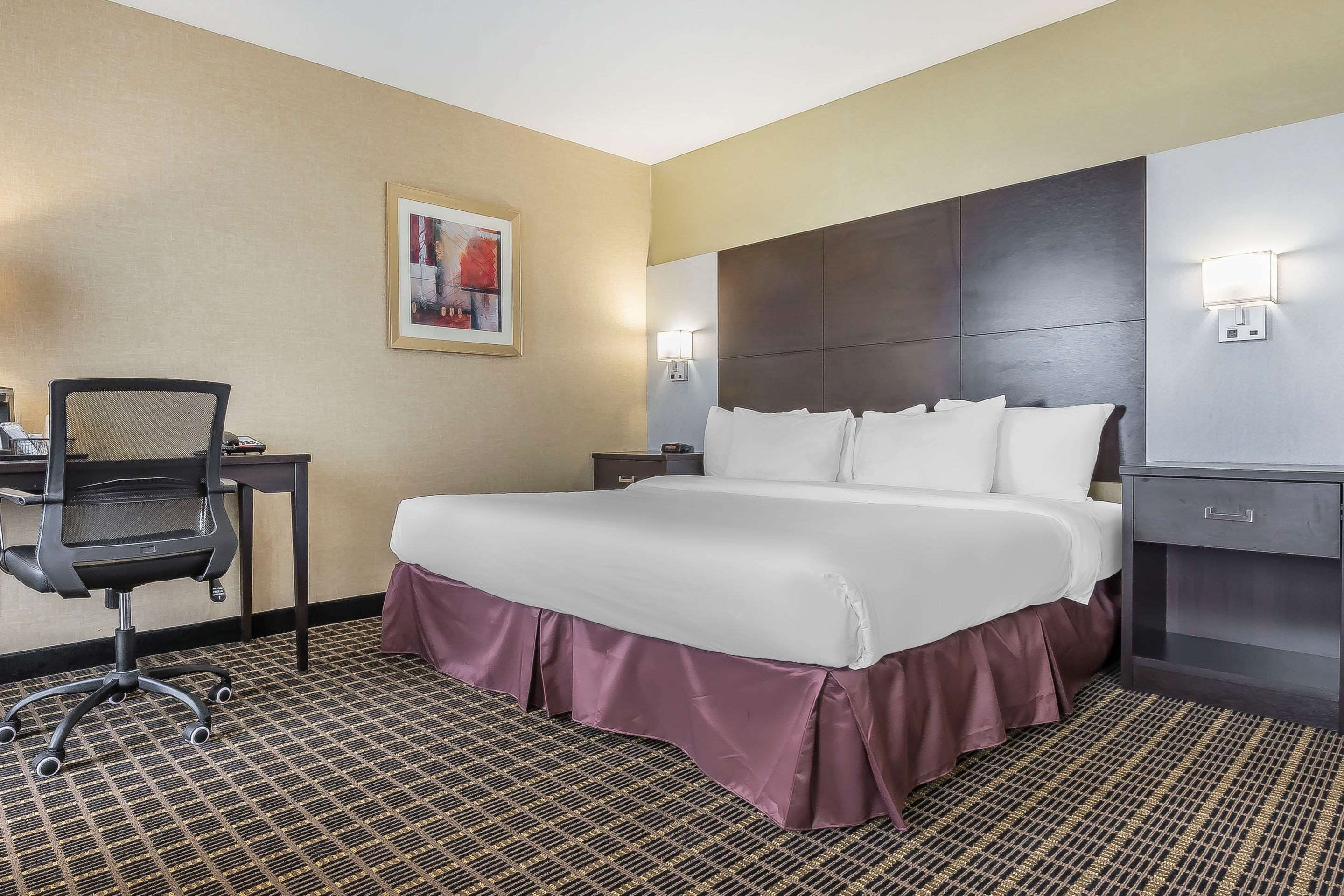 Habitación de hotel con una cama grande, un escritorio con silla, una mesita de noche y una lámpara de pared.