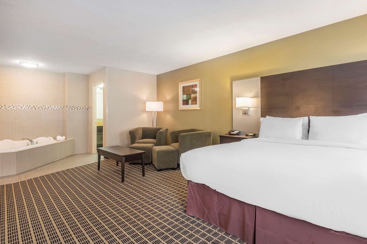 A hotel room with a large bed, a desk, two armchairs, a sofa, a bathroom with a bathtub, and a patterned carpet.