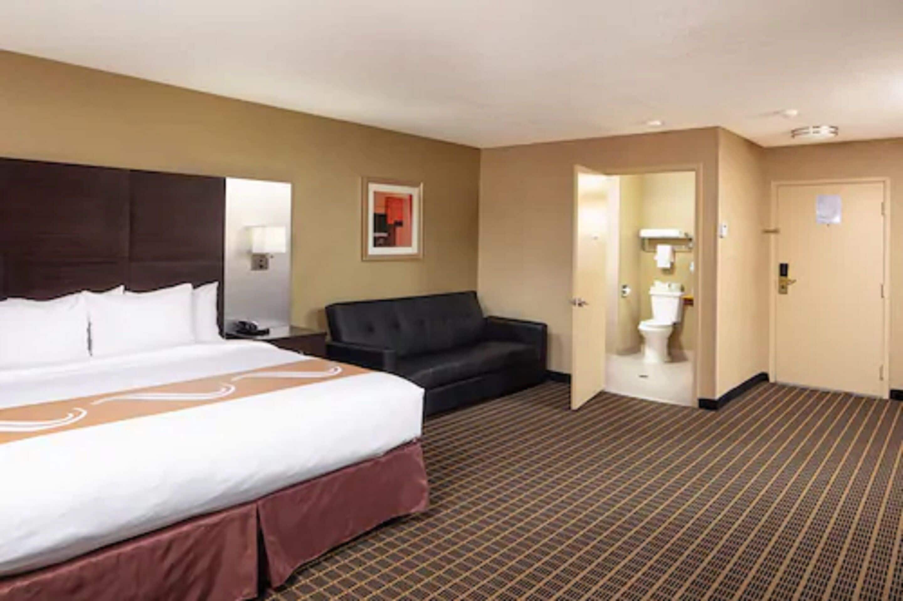 Quarto de hotel com uma cama grande, um sofá preto, uma mesa pequena e um banheiro visível através de uma porta aberta.
