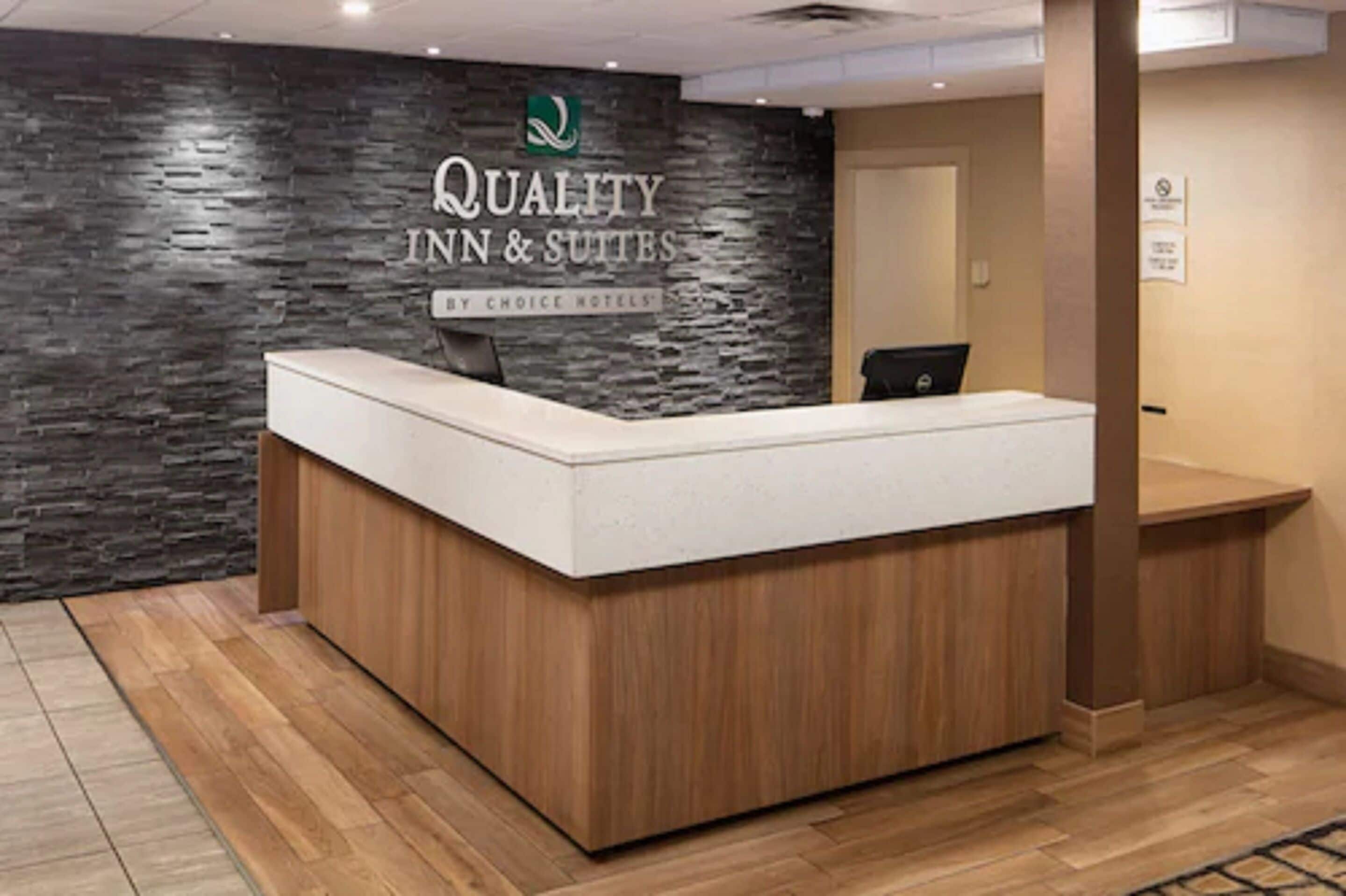 Balcão de recepção com parede de pedra ao fundo e placa com a inscrição "Quality Inn & Suites by Choice Hotels".