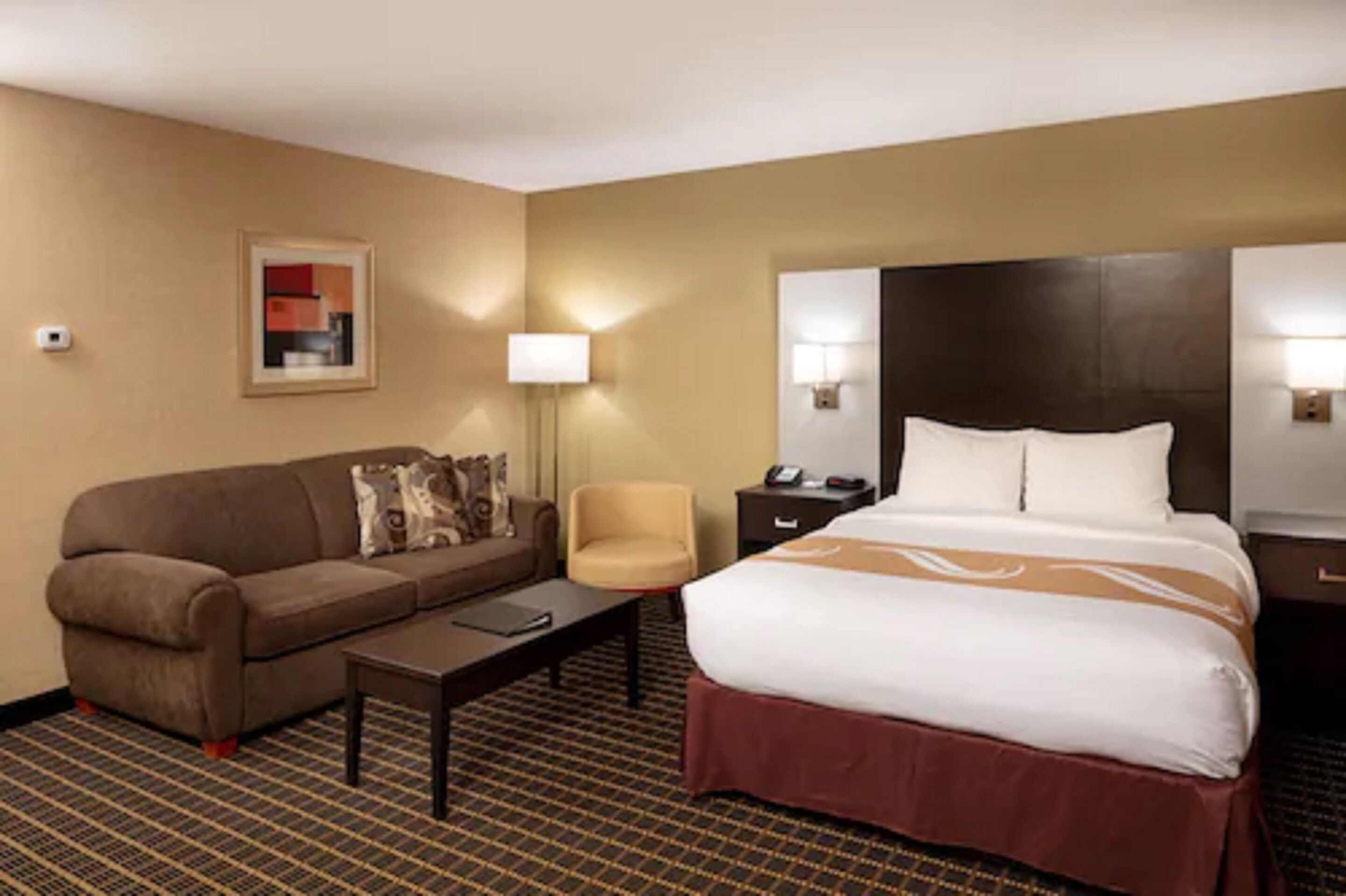 Une chambre d’hôtel avec un lit, un canapé, une chaise, une table de chevet et une lampe.