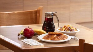 Una mesa de madera con alimentos para el desayuno, incluyendo gofres, un tazón de cereales, una botella de sirope, un plátano y una manzana.