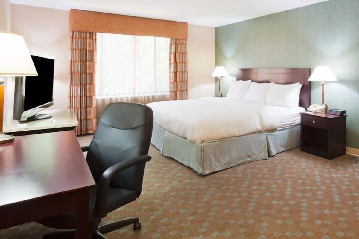 A hotel room with a bed, two bedside lamps, a desk with a computer, a chair, and a window with curtains.