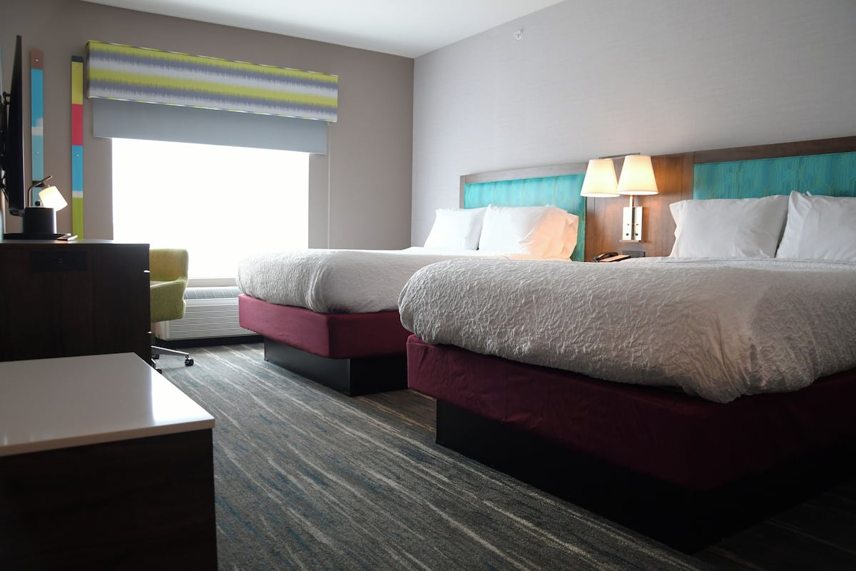 A hotel room with two beds, a desk, a chair, and a large window with blinds.