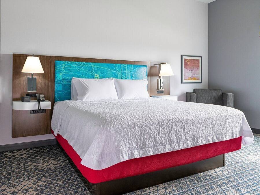 Habitación de hotel con una cama grande, dos mesitas de noche, cabecera con diseño de paisaje urbano y un detalle rojo en la cama, además de un piso con estampado.