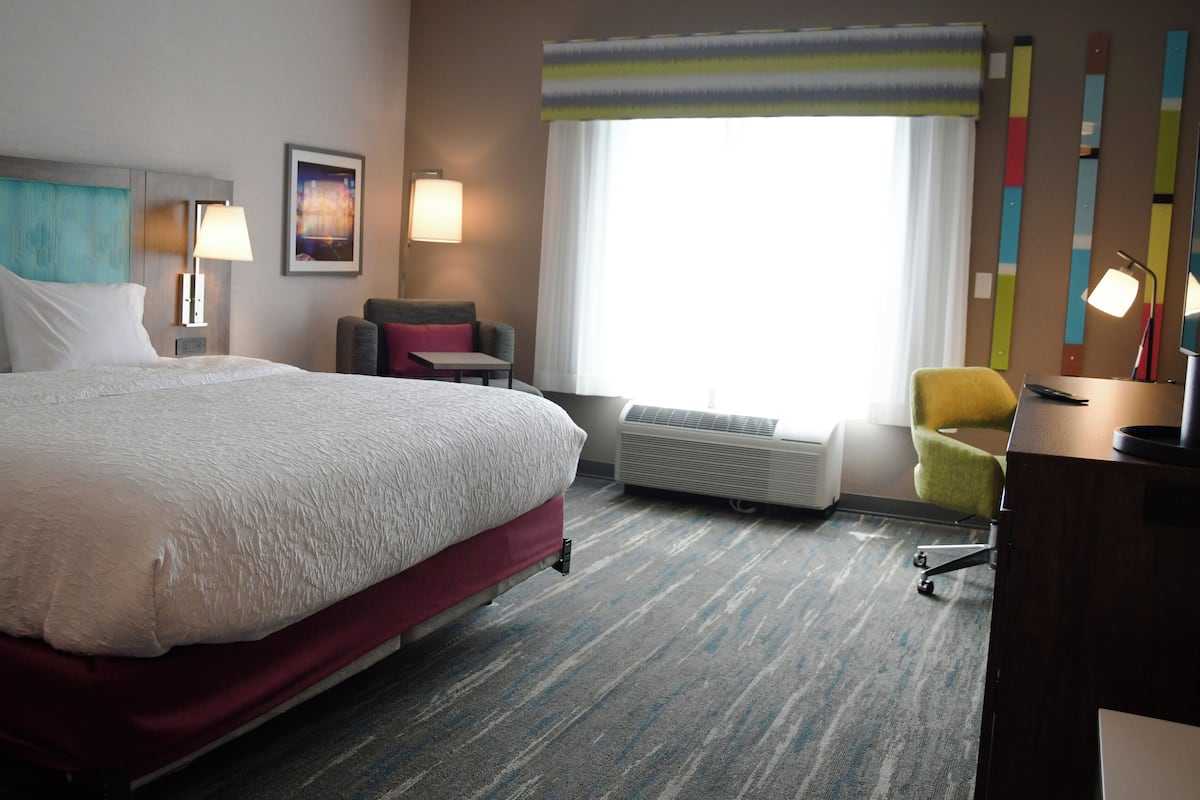 A hotel room with a large bed, a desk, a chair, a window with curtains, and a wall with colorful decorations.