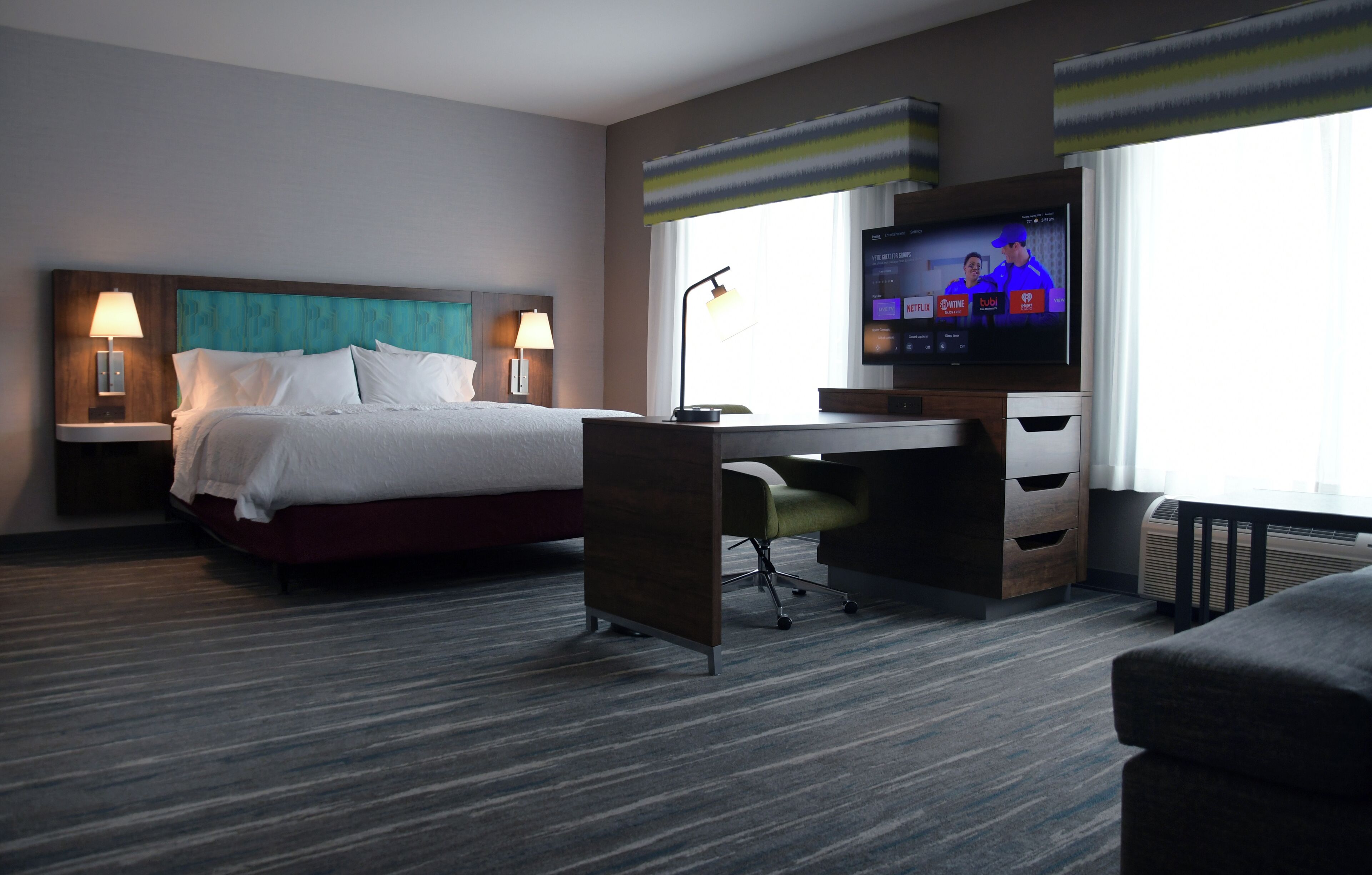 Habitación de hotel con cama, escritorio, silla, televisor y ventanas amplias.
