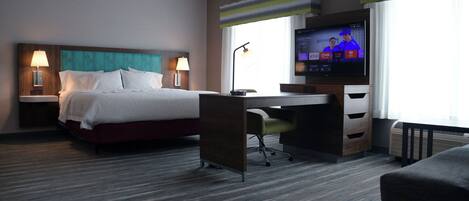 Habitación de hotel con cama, escritorio, silla, televisor y ventanas amplias.