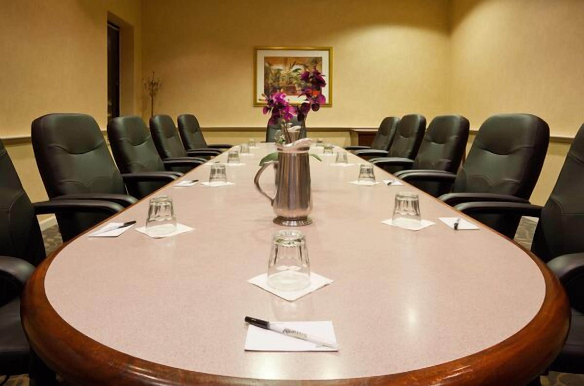 A conference room with a long table, black chairs, a vase with flowers, and a pitcher.