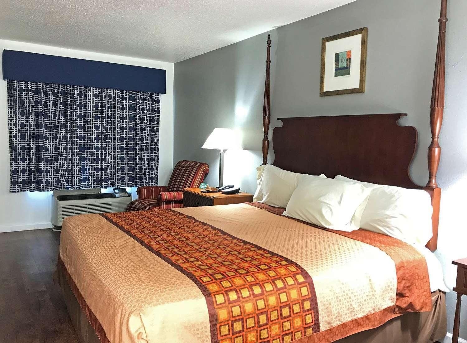 Habitación de hotel con una cama con dosel, pared con estampado, una silla, un escritorio con lámpara y un cuadro en la pared.