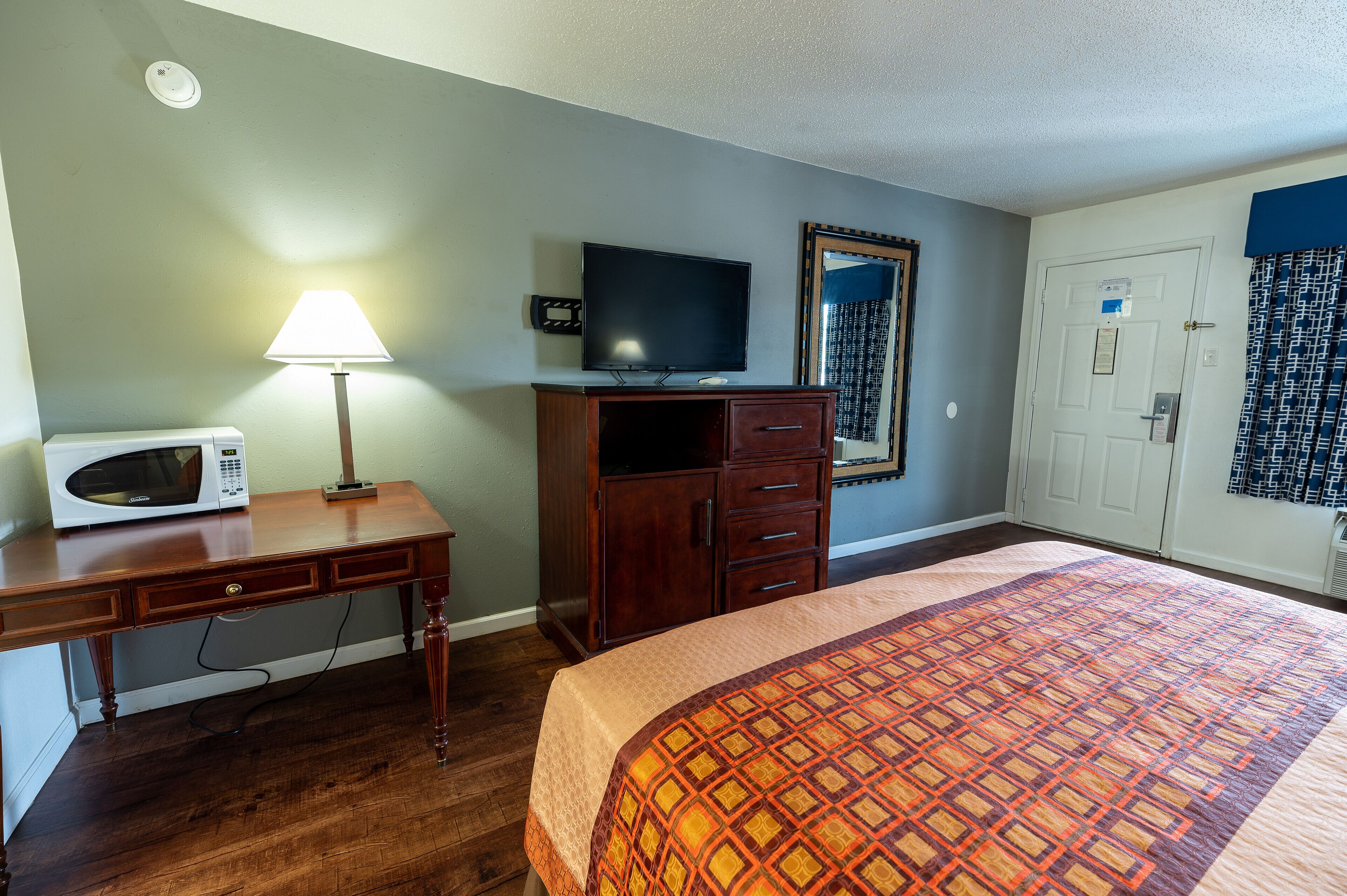 A hotel room with a bed, a desk with a lamp, a television, a microwave, a wardrobe, and a door with a mirror.