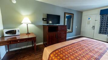 A hotel room with a bed, a desk with a lamp, a television, a microwave, a wardrobe, and a door with a mirror.