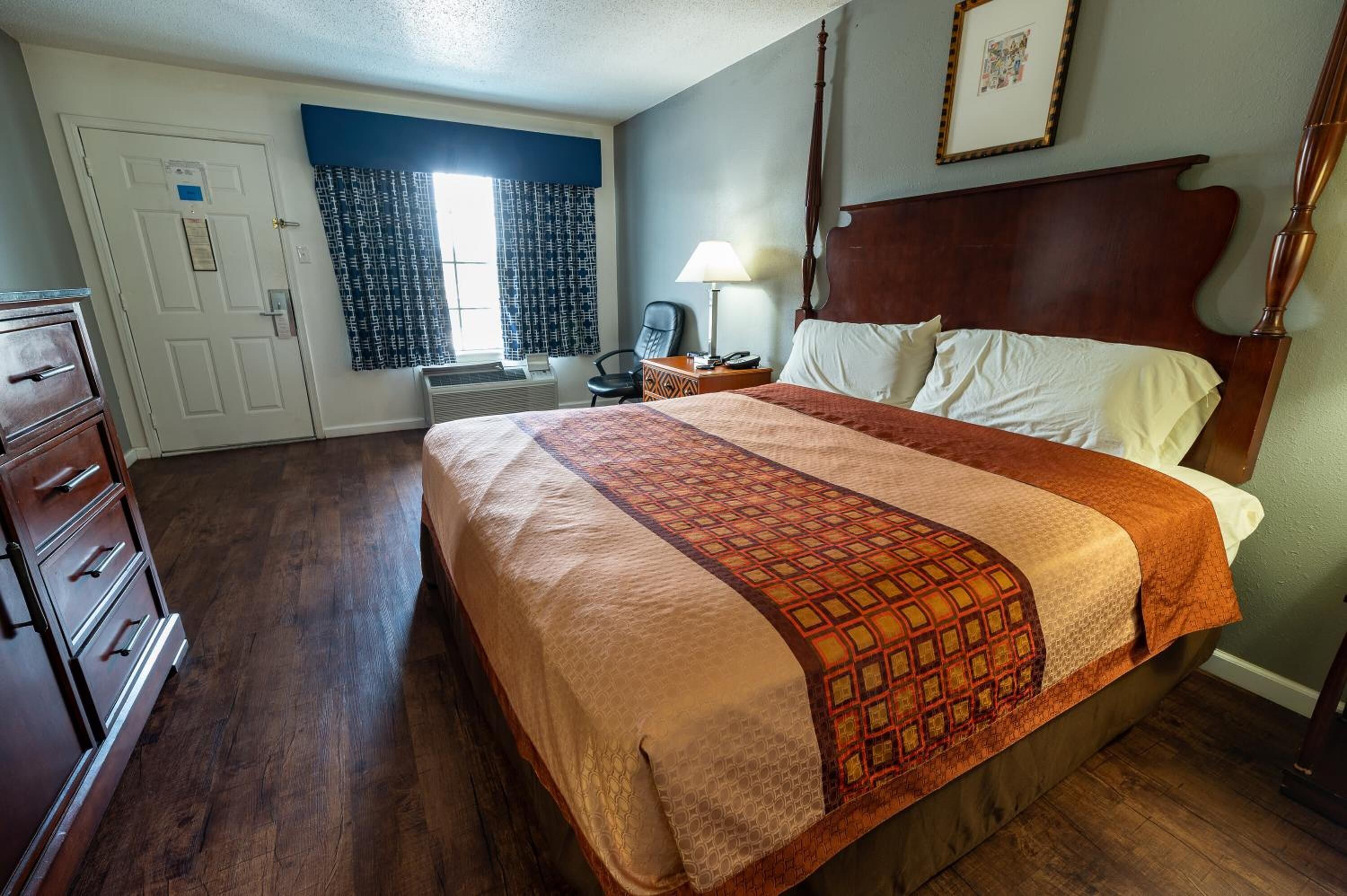 Una habitación de hotel con una cama grande, un aparador de madera, un escritorio con lámpara y una ventana con cortinas estampadas.