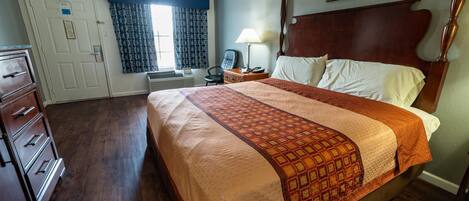 Una habitación de hotel con una cama grande, un aparador de madera, un escritorio con lámpara y una ventana con cortinas estampadas.
