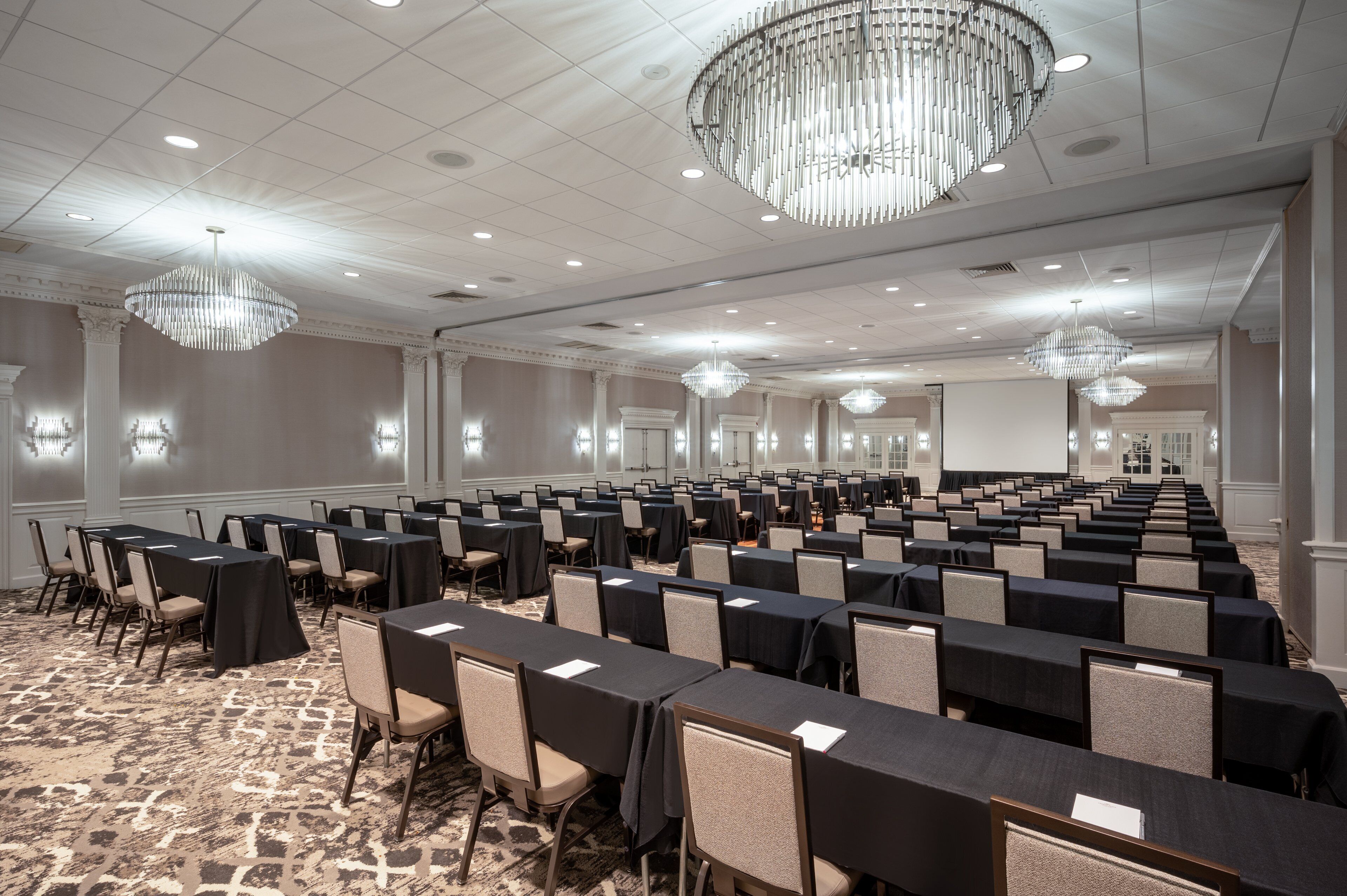 A large conference room with rows of tables and chairs, chandeliers, and a carpeted floor.