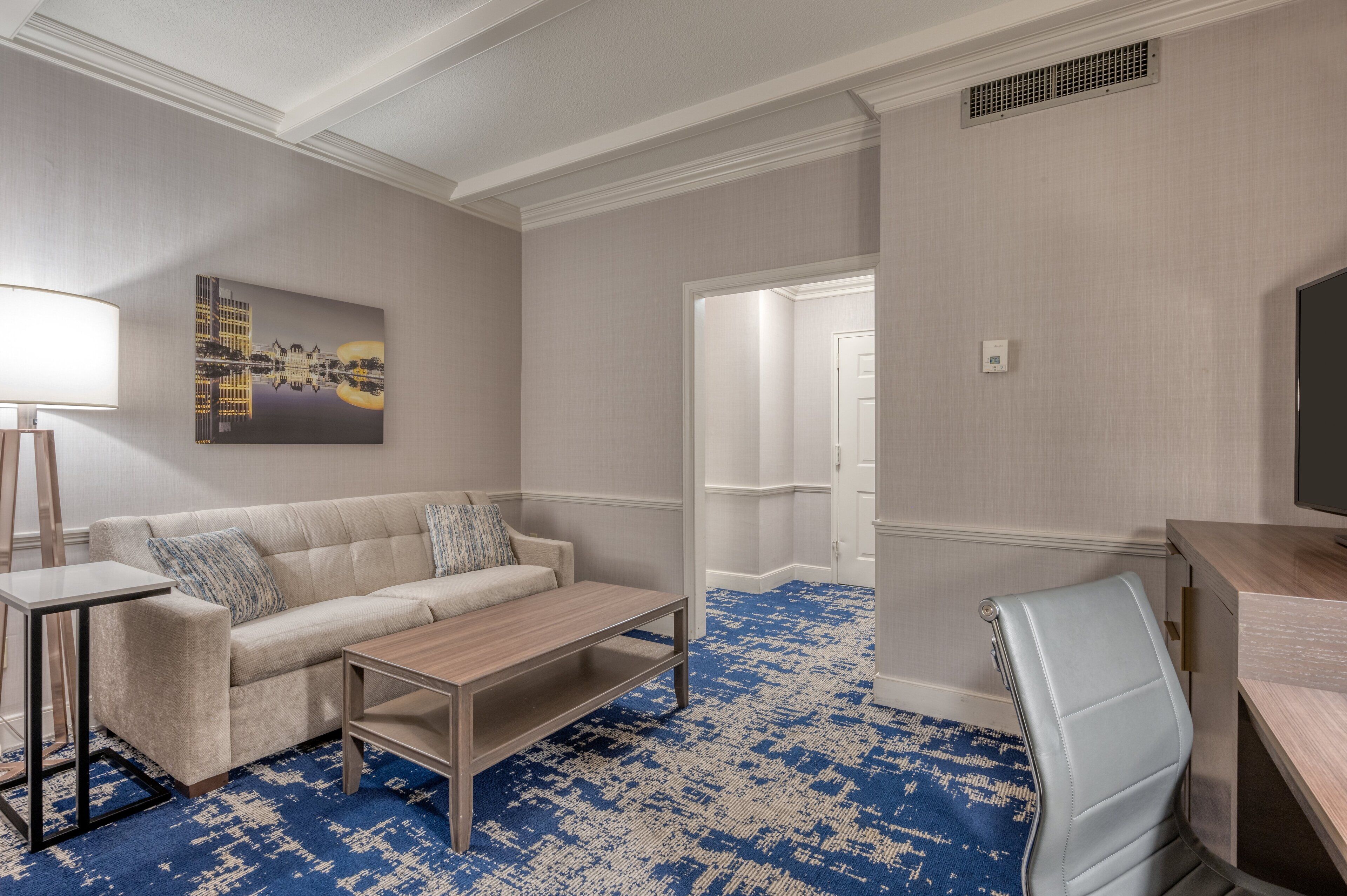 A hotel room with a beige sofa, a wooden coffee table, a blue patterned carpet, a desk with a chair, and a painting on the wall.