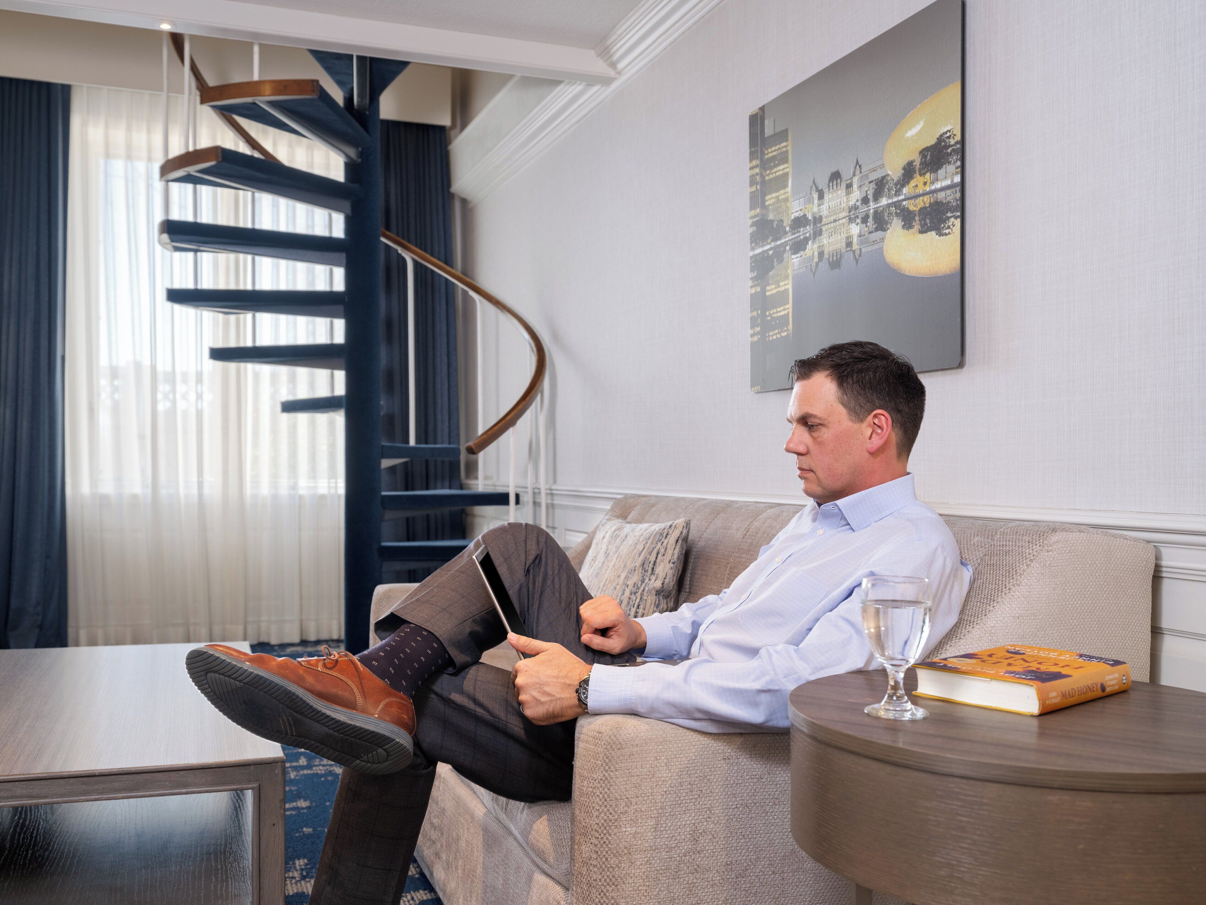 A man sitting on a sofa in a well-lit room with a modern staircase, a cityscape painting on the wall, and a book on a side table.