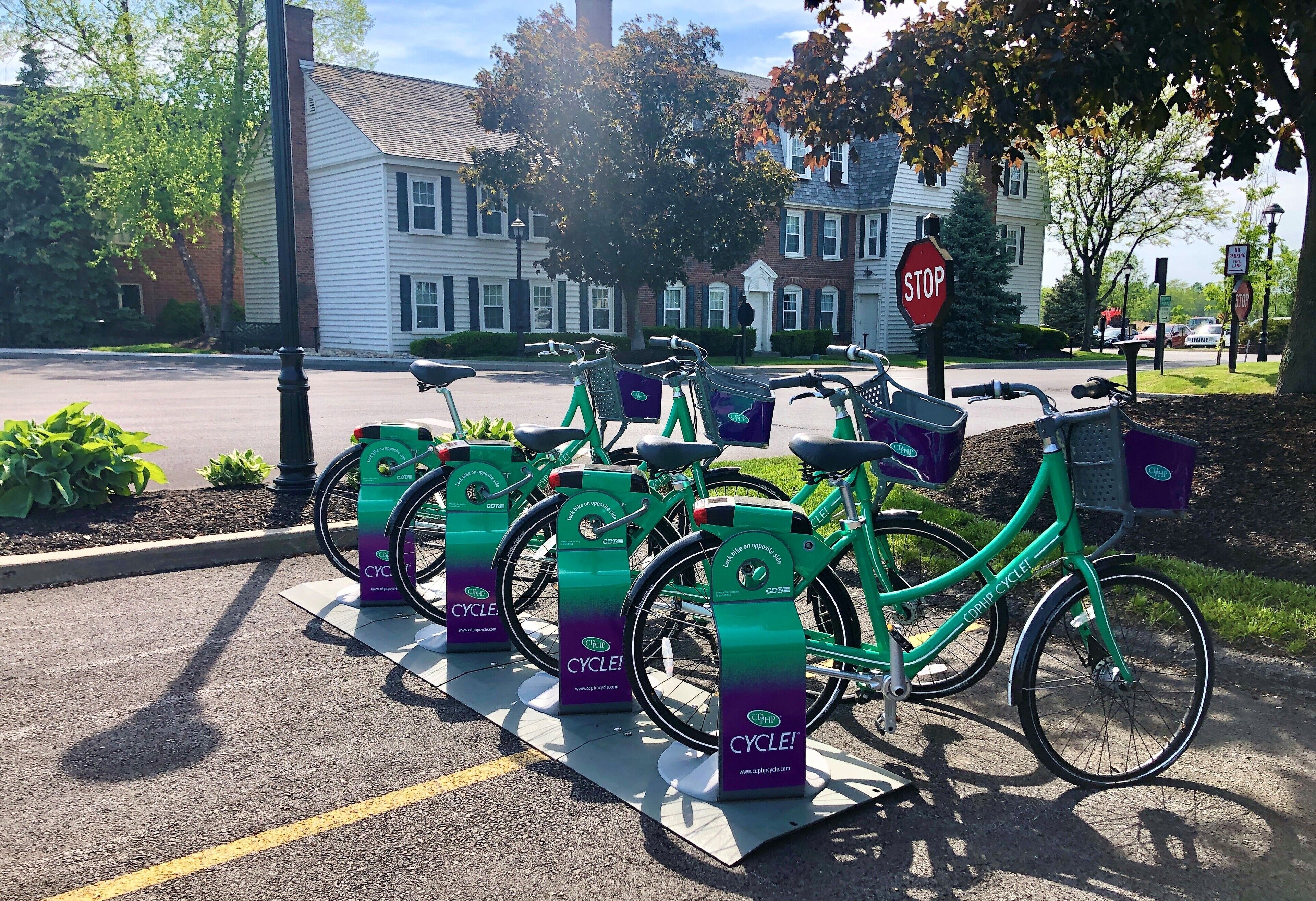 A row of green bicycles parked at a bike-sharing station with a stop sign and residential buildings in the background.