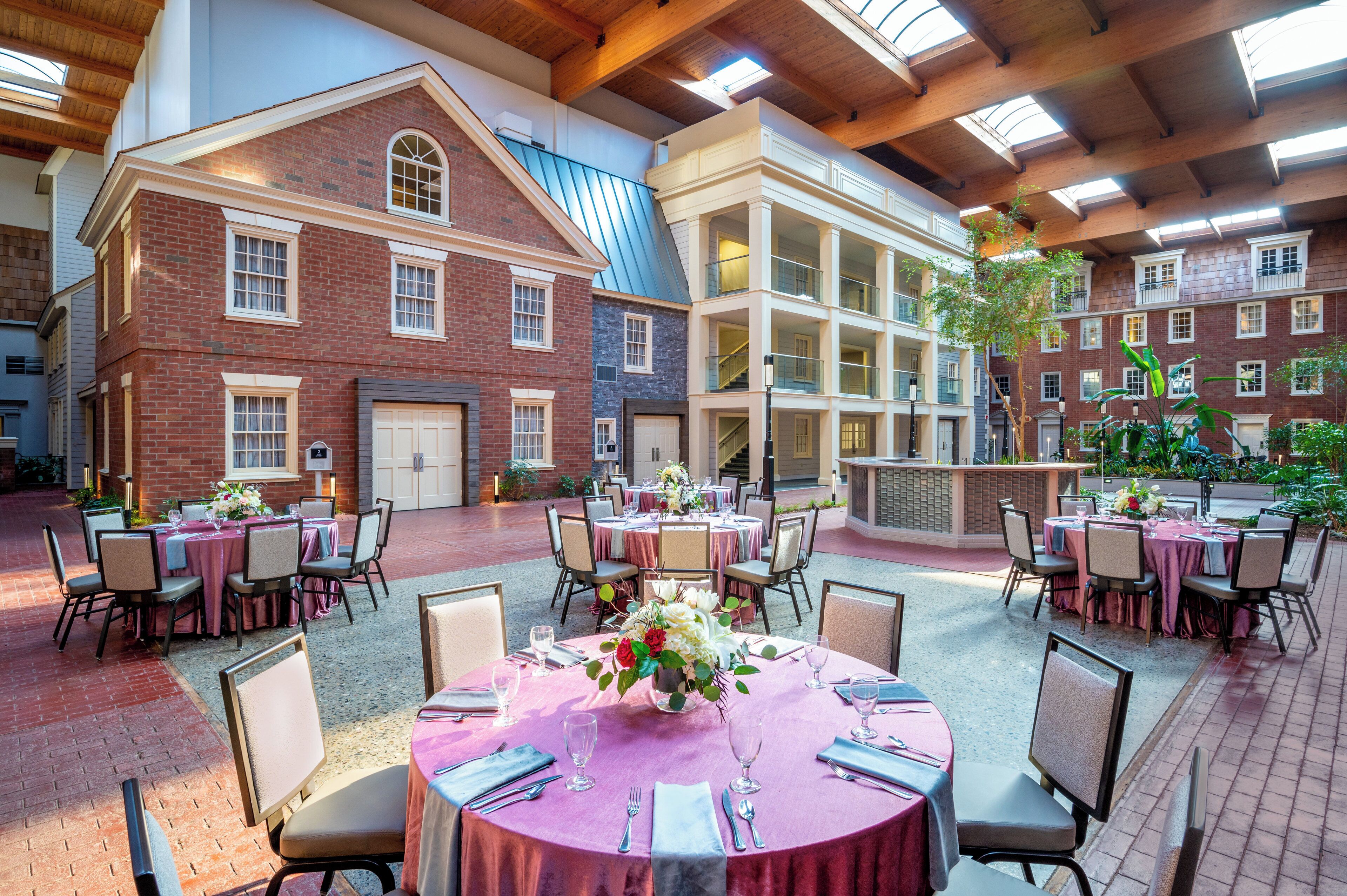 A spacious indoor dining area with multiple tables set for guests, surrounded by brick buildings with large windows and a high ceiling with wooden beams.