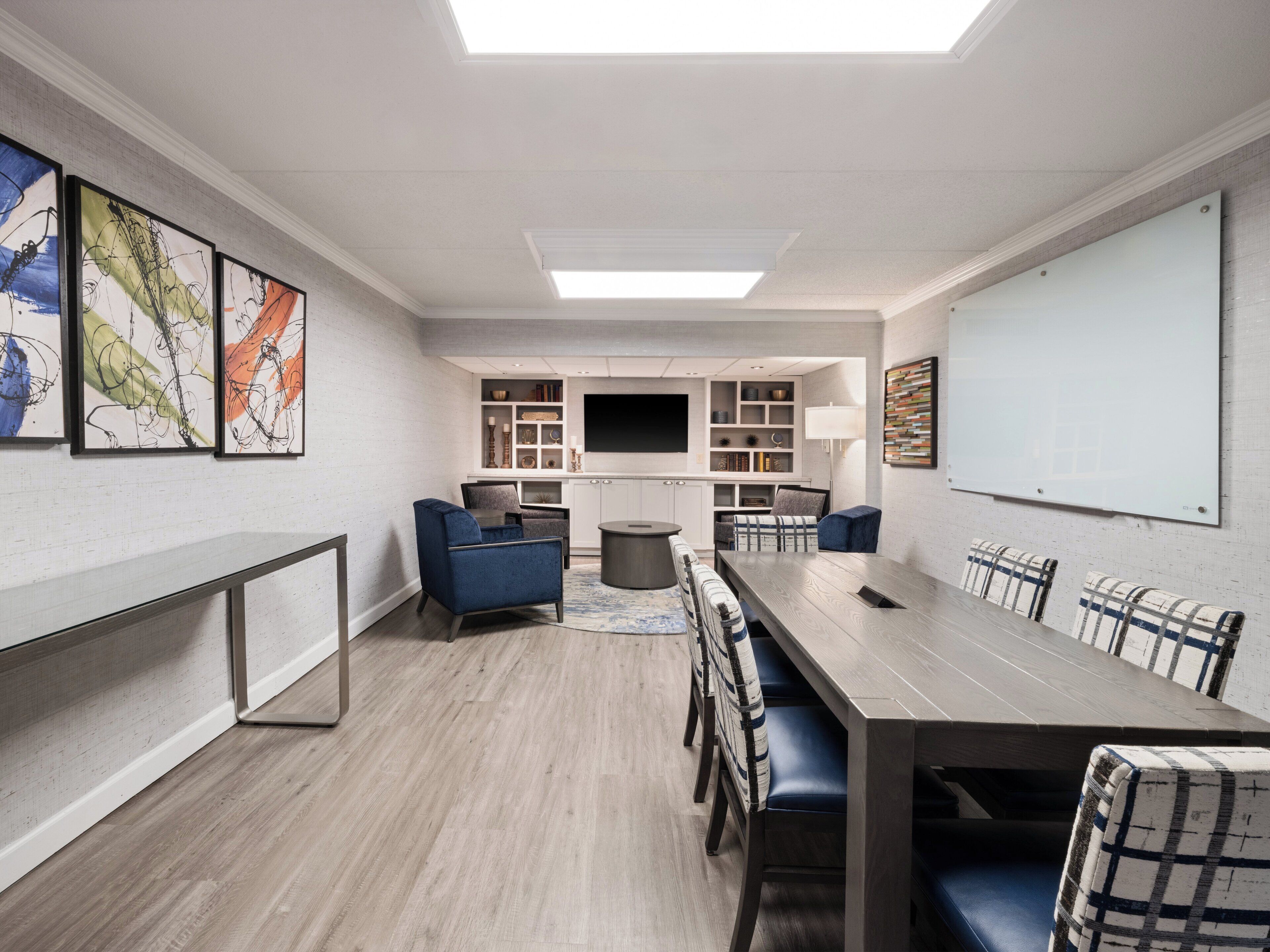 A modern conference room with a long table, blue and plaid chairs, a whiteboard, and abstract art on the walls.
