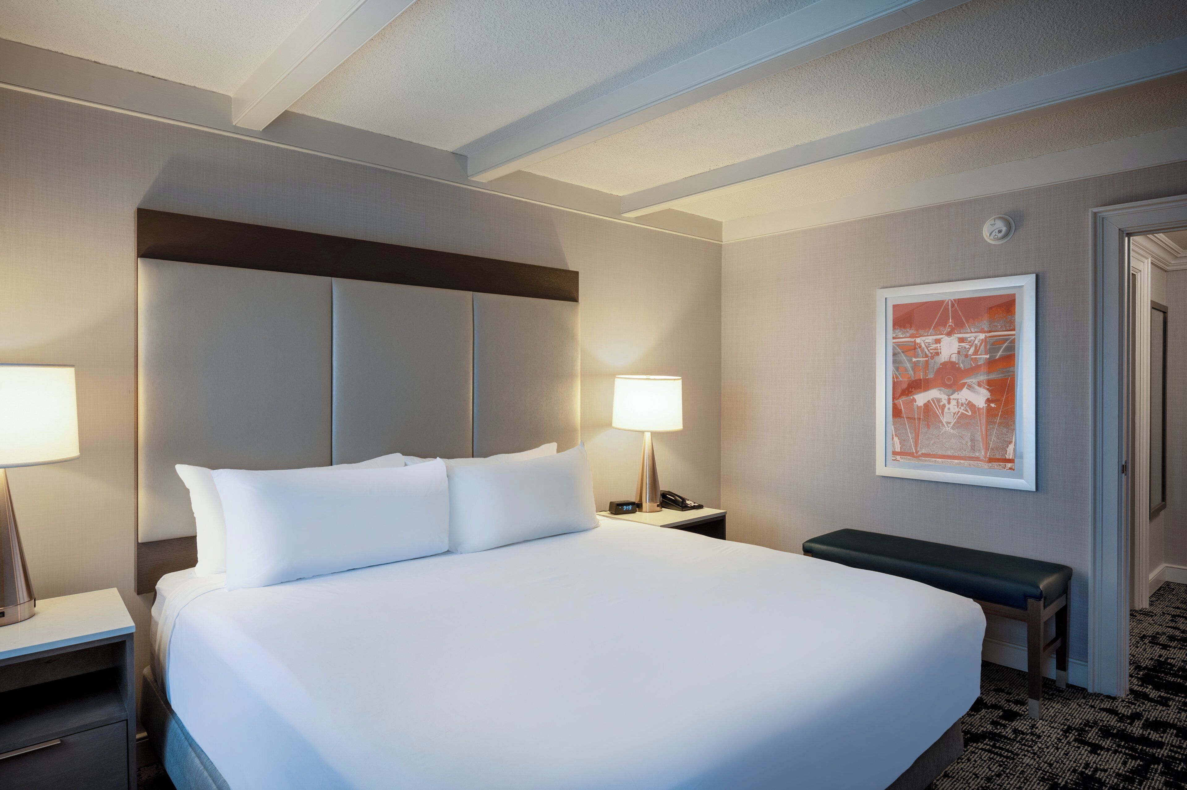 Una habitación de hotel con una cama grande, lámparas de noche, una mesita de noche, un cuadro enmarcado en la pared y un banco.